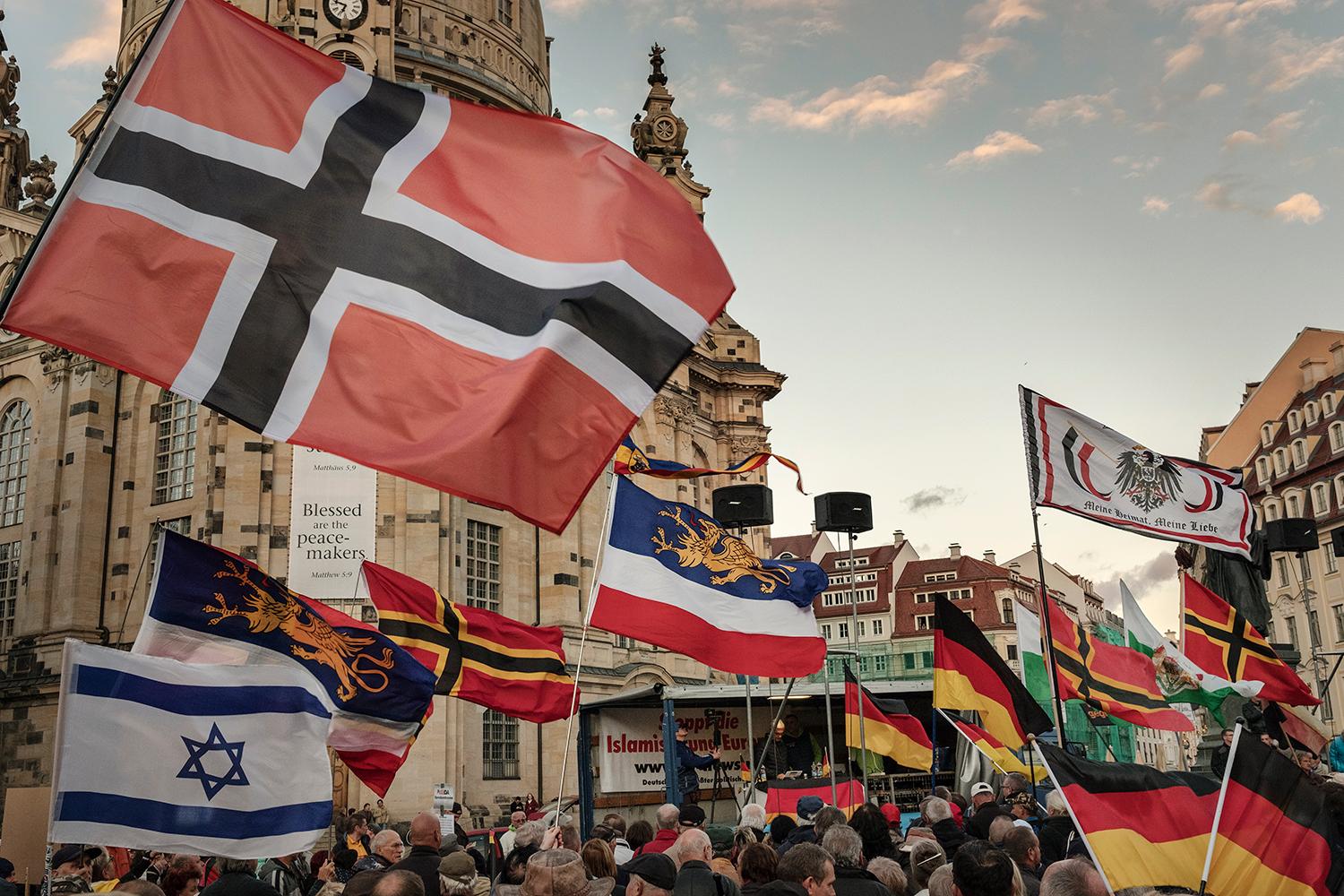 Demonštranti mávajú vlajkami rôznych štátov a nemeckých krajov. Na pódiu rečnia čelní predstavitelia nacionalistického hnutia Pegida.