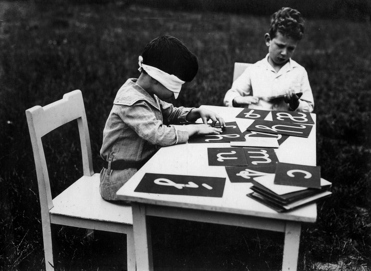 Deti si v Montessori škôlke precvičujú znalosť abecedy. Tvary vyrezaných písmen sa usilujú rozoznávať predovšetkým hmatom, niekedy aj so zaviazanými očami. Fotografia spolu s ďalšími dvomi je z Nemecka z roku 1925.